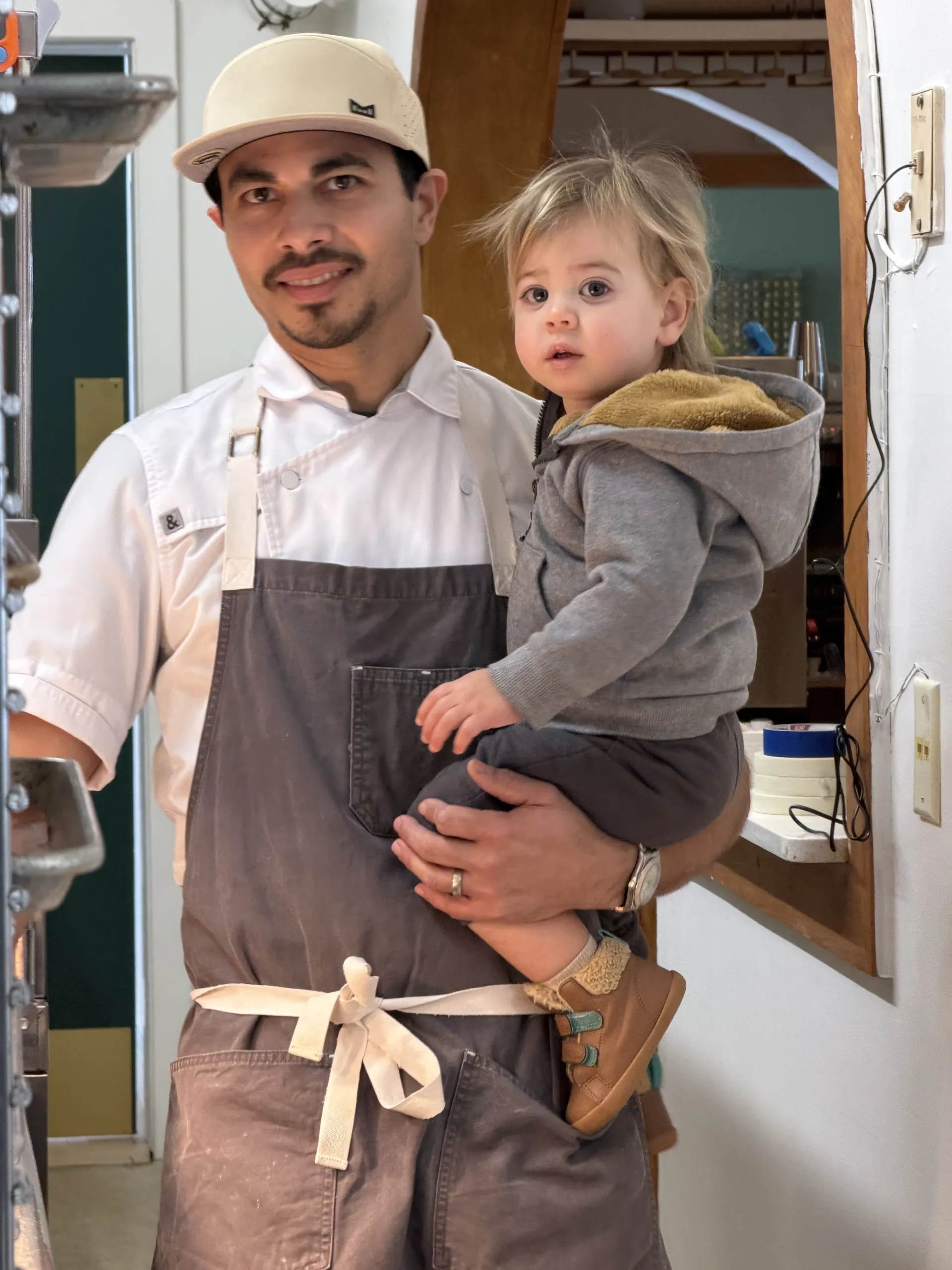 Chef Miguel Calderon in his whites, holding his youngest daughter in the Alder kitchen