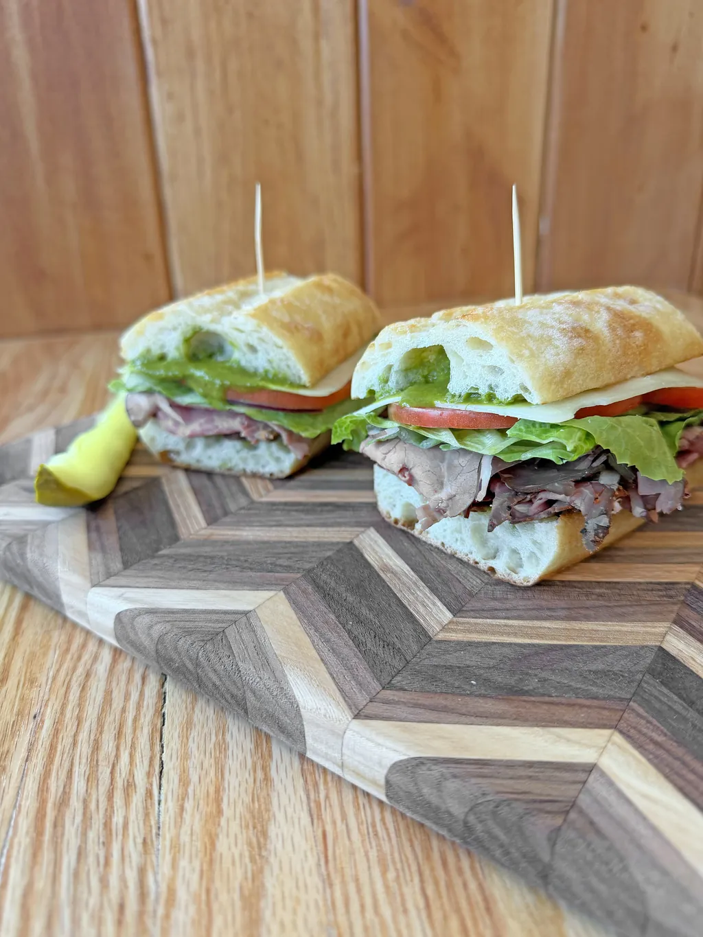 Roast beef sandwich on ciabatta with cheddar, lettuce, and tomato, cut in half and served on a wooden board with a pickle spear